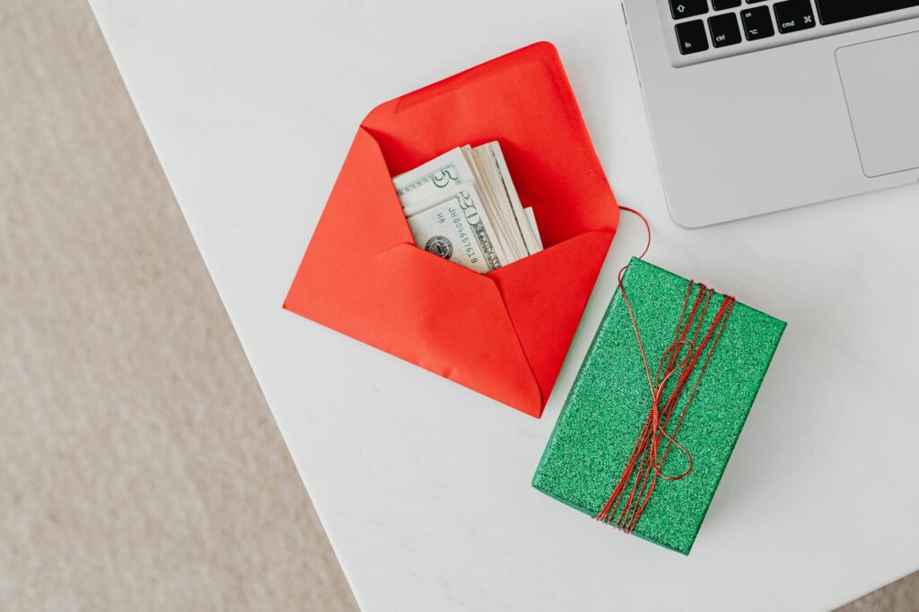 A green gift box and red envelope with cash on a desk beside a laptop, emphasizing holiday or financial themes.